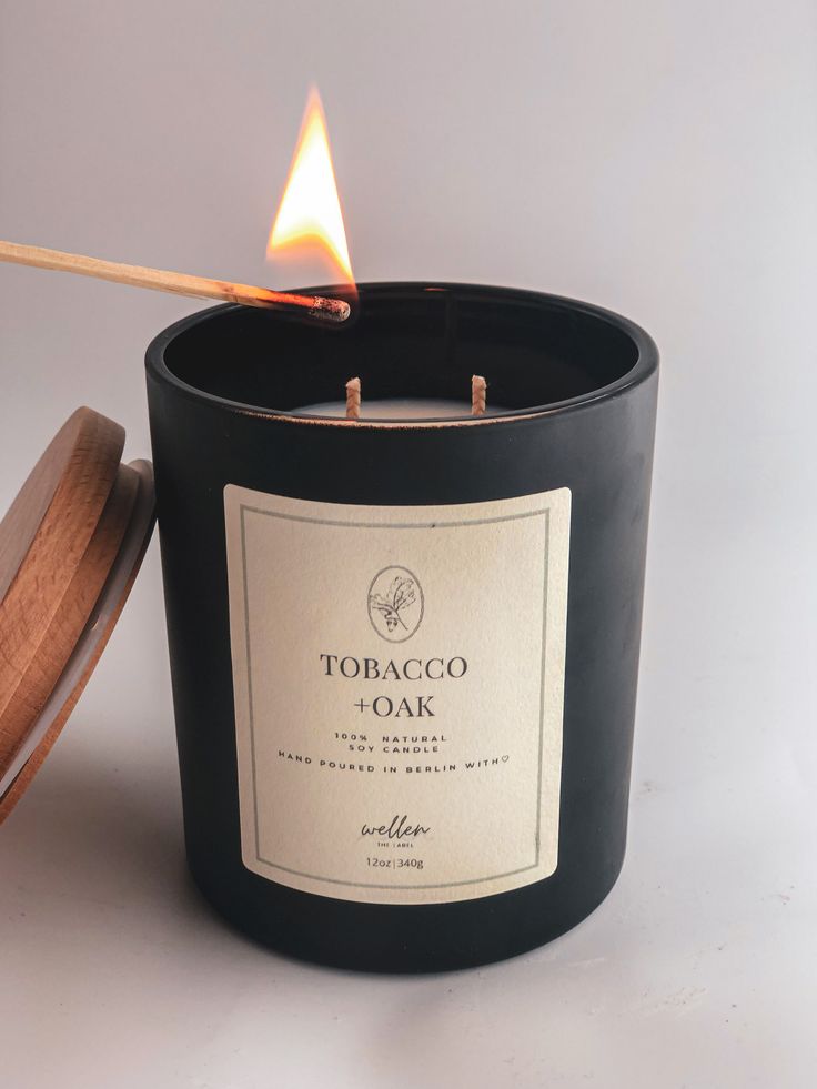 a black candle with a wooden lid next to a lit matchstick on a white surface