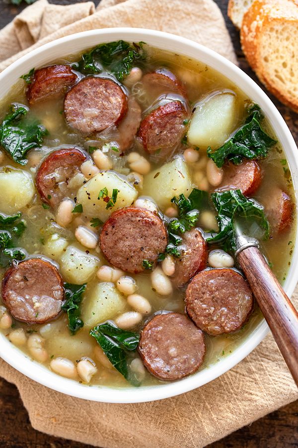 a white bowl filled with sausage, beans and spinach next to bread on a wooden table