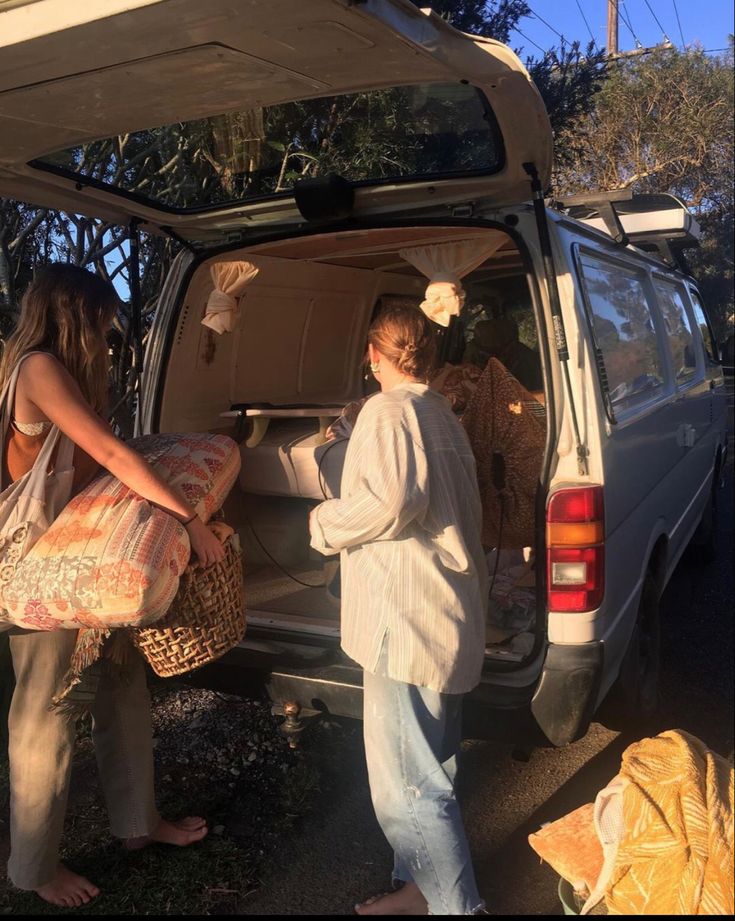 two women loading items into the back of a van with their trunk open, while another woman looks on