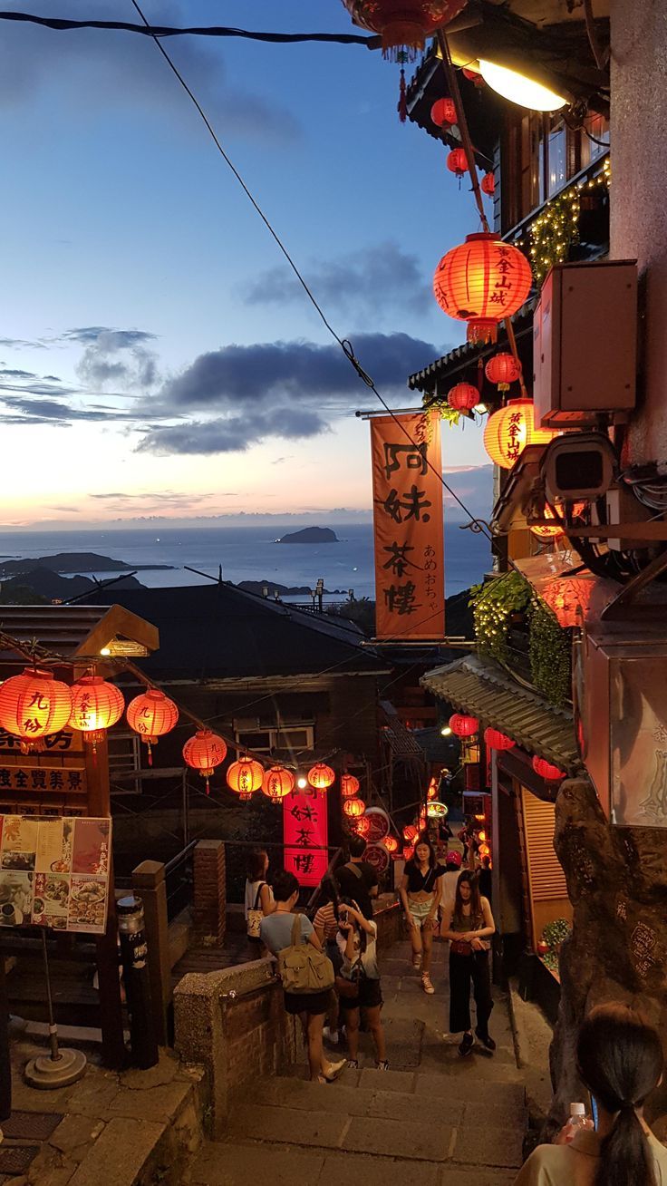 many people are walking down the street with lanterns hanging from the side of buildings at night