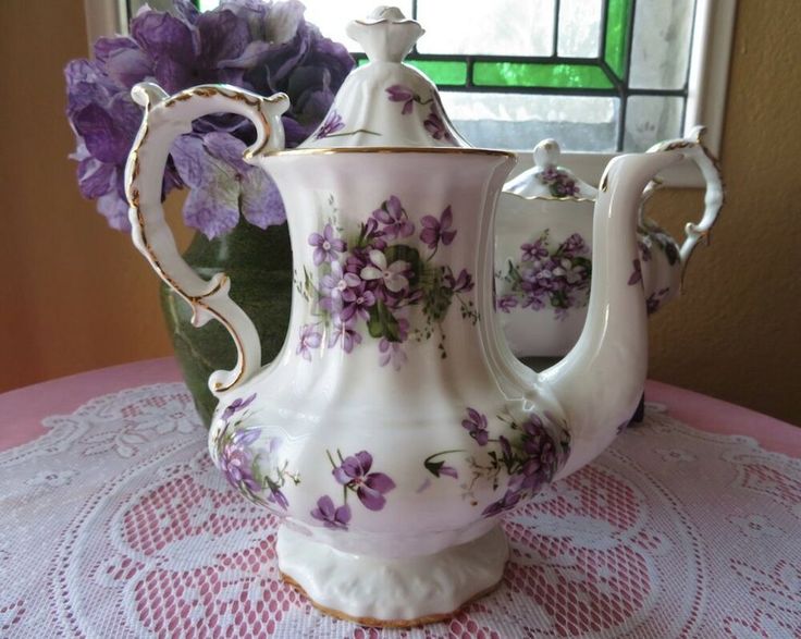 a white tea pot sitting on top of a table next to a vase filled with purple flowers