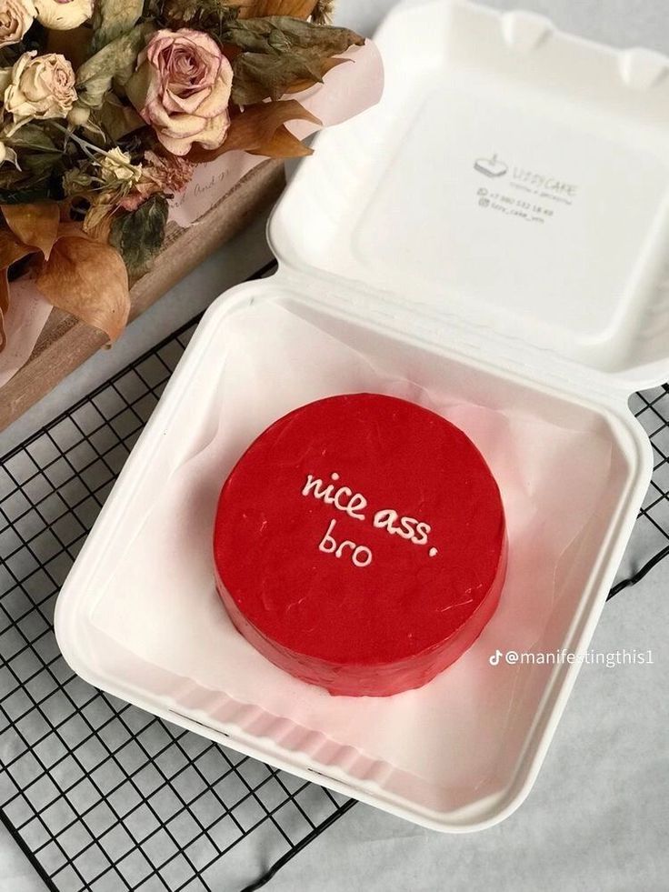 a red piece of cake sitting in a white container on a table next to flowers