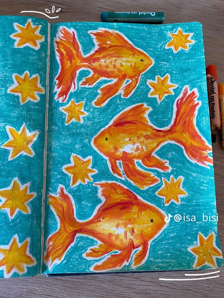 an open notebook with goldfish and stars on it, next to crayon markers