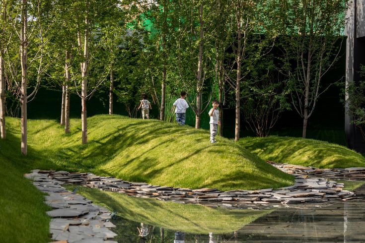 two people walking down a grassy hill next to a pond and forest filled with rocks