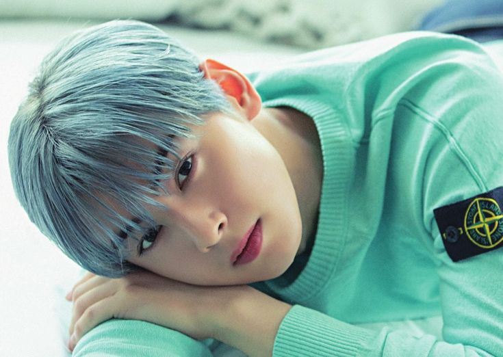 a young boy with grey hair wearing a green shirt and sitting on a white bed