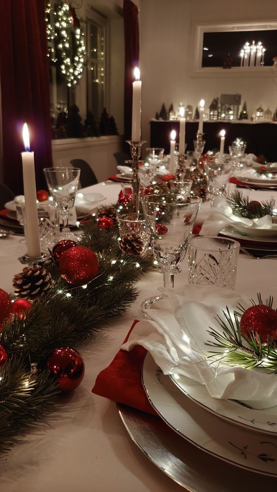 the table is set for christmas dinner with silverware and red ornaments on it, along with candles