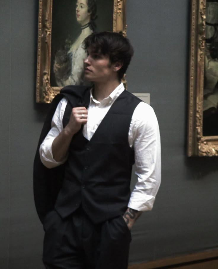 a man standing in front of two paintings wearing a black vest and white dress shirt