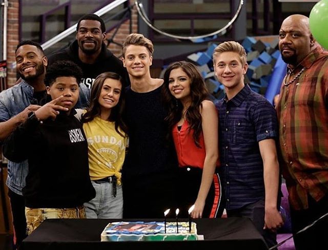 a group of people standing around a table with a cake in front of them and balloons