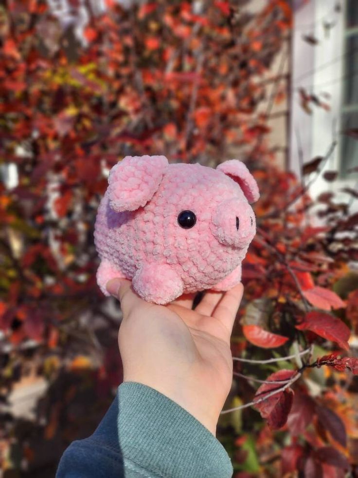 a hand holding a pink stuffed animal in front of some trees with red leaves on it