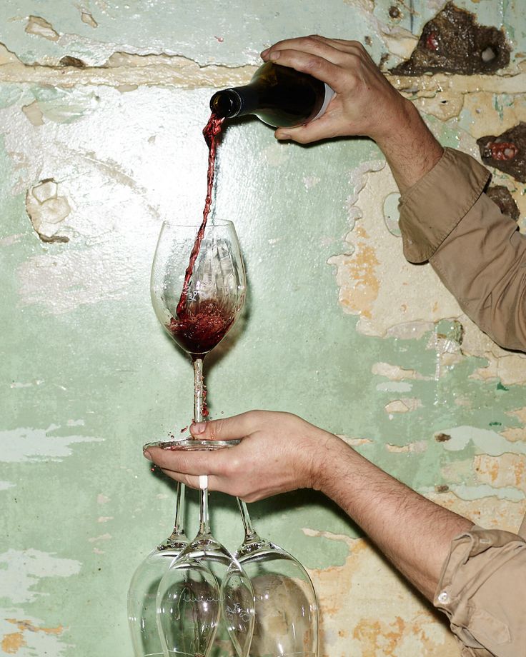 a man pours red wine into two wine glasses in front of a peeling green wall