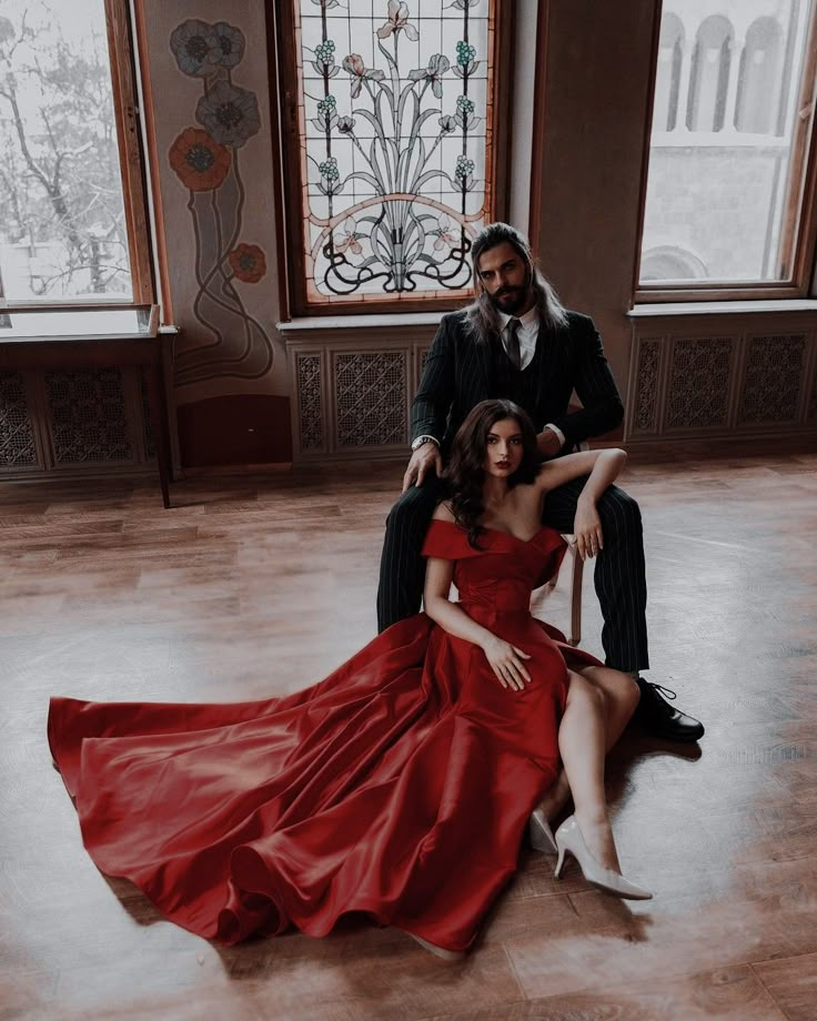 a man and woman sitting on the floor in front of two windows wearing red dresses