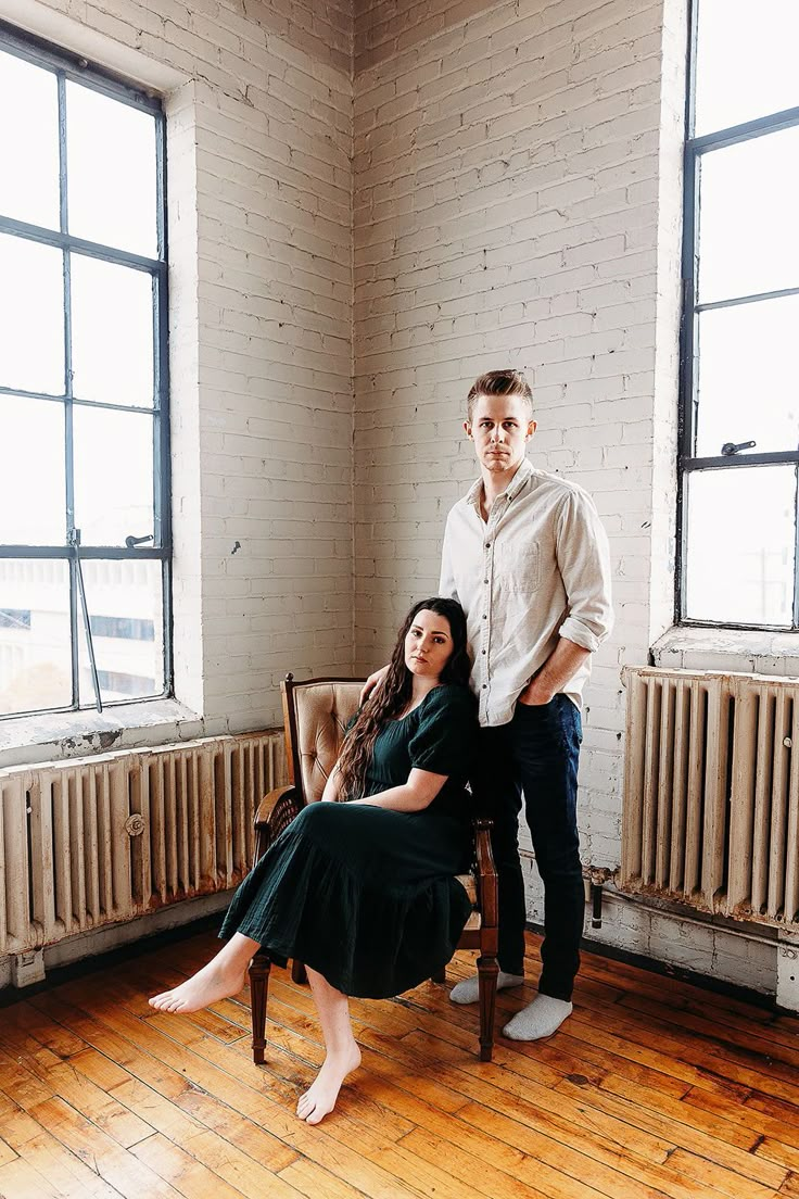 a man and woman sitting on a chair in an empty room next to two windows