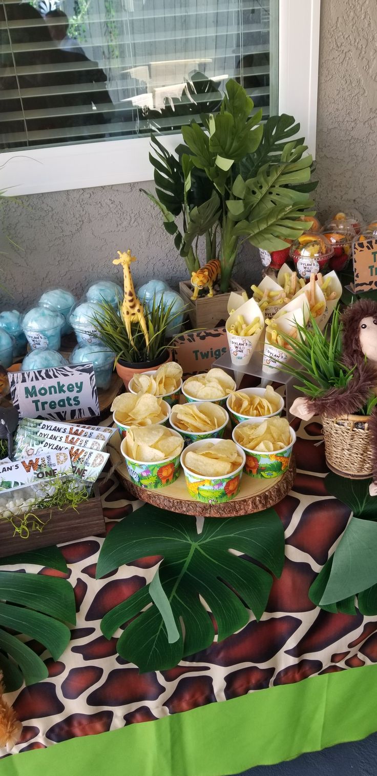 a table topped with lots of food next to a window filled with plants and stuffed animals