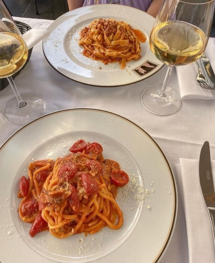 two plates of spaghetti with tomato sauce and glasses of wine on the table in front of them