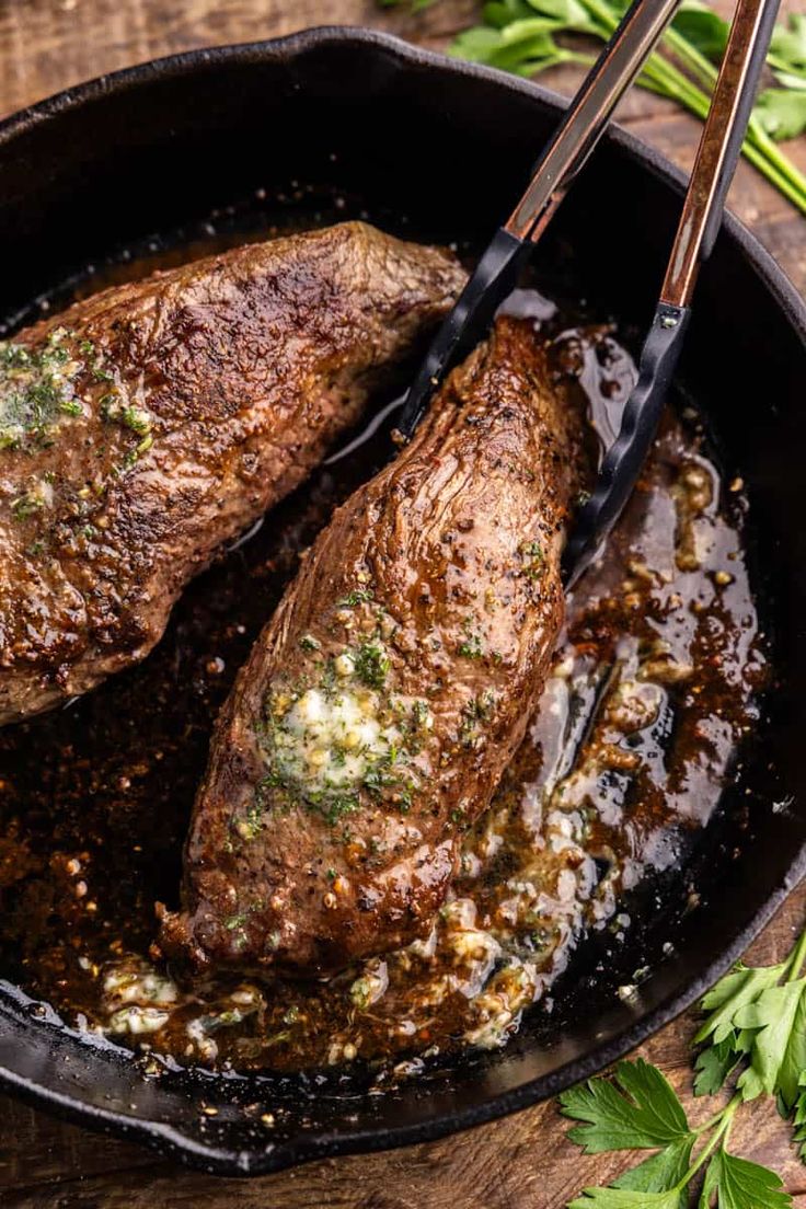 two steaks are being cooked in a skillet with sauce and herbs on the side