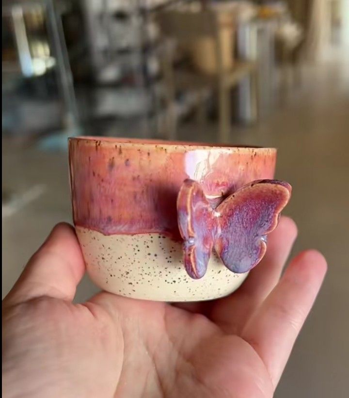 a hand holding a small ceramic cup with a butterfly on the top and bottom, in front of a blurry background