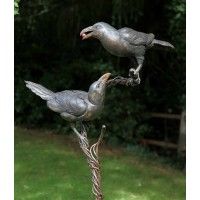 two metal birds perched on top of a tree branch in the grass with trees behind them