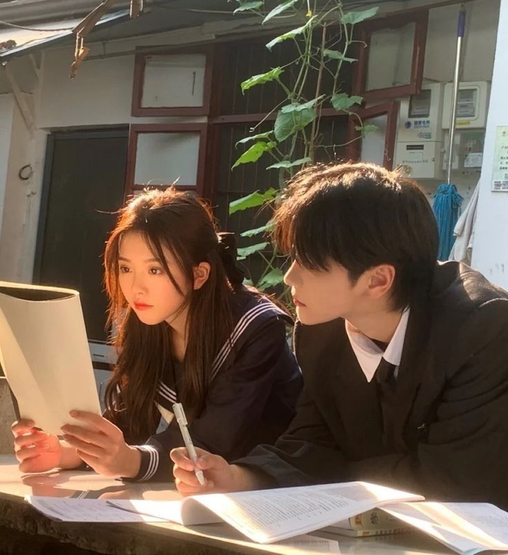 two people sitting at a table with papers and pens in front of them, looking at something on the computer screen