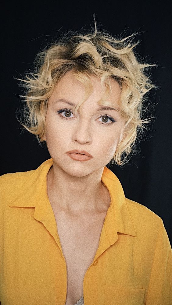 a woman with blonde hair wearing a yellow shirt and looking at the camera while standing in front of a black background