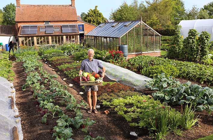 an older man holding a basket full of vegetables in a garden with greenhouses behind him