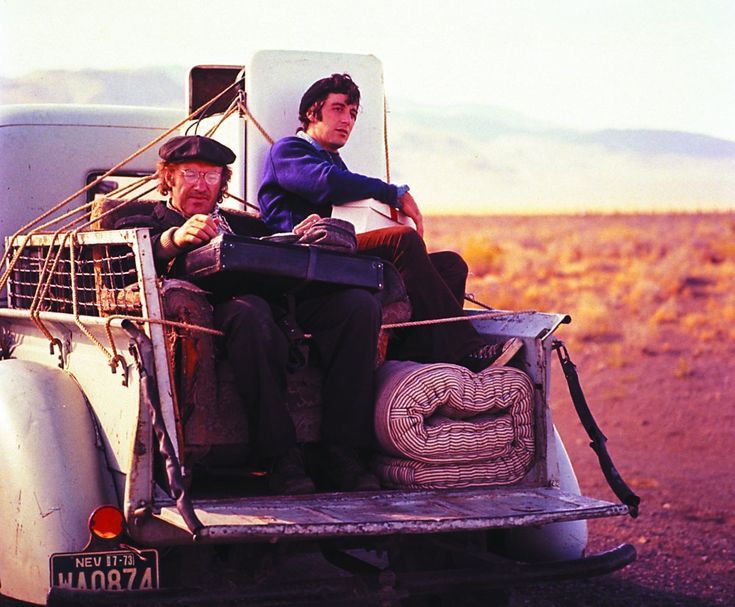 two men sitting in the back of an old pick up truck with luggage strapped to it