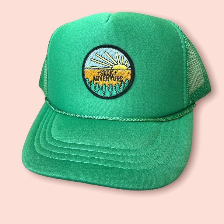 a green trucker hat with an image of the sun and trees on it, against a pink background