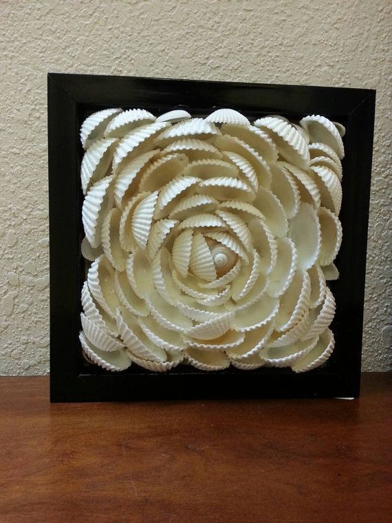 a white flower made out of shells in a black frame on top of a wooden table