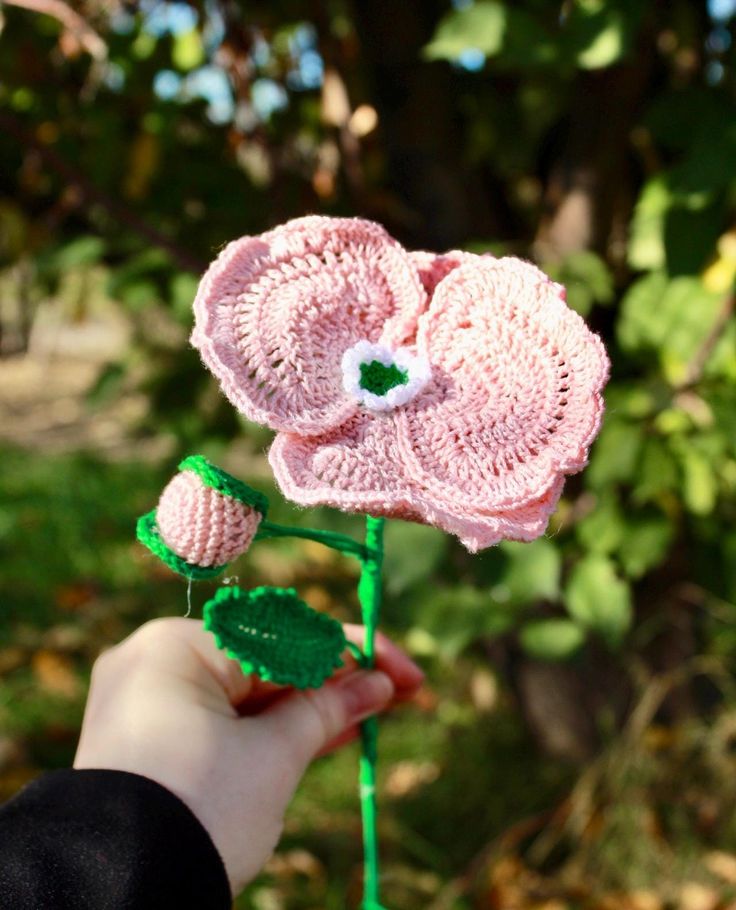 a hand holding a crocheted pink flower with green stems and leaves on it