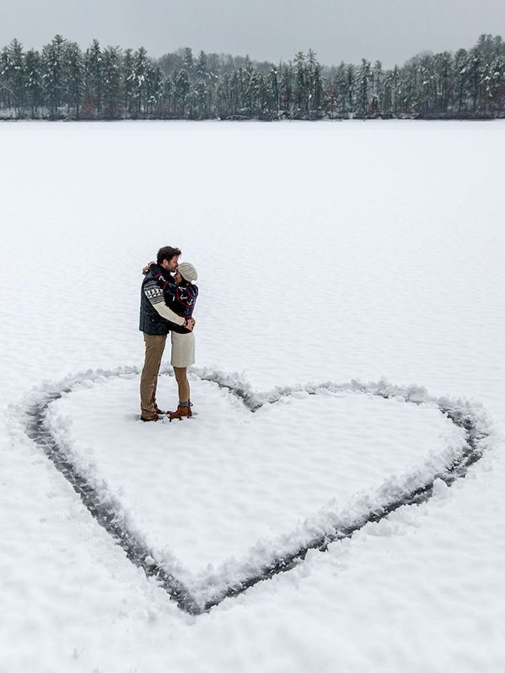 a man and woman standing in the middle of a heart shaped snow covered field with their arms around each other