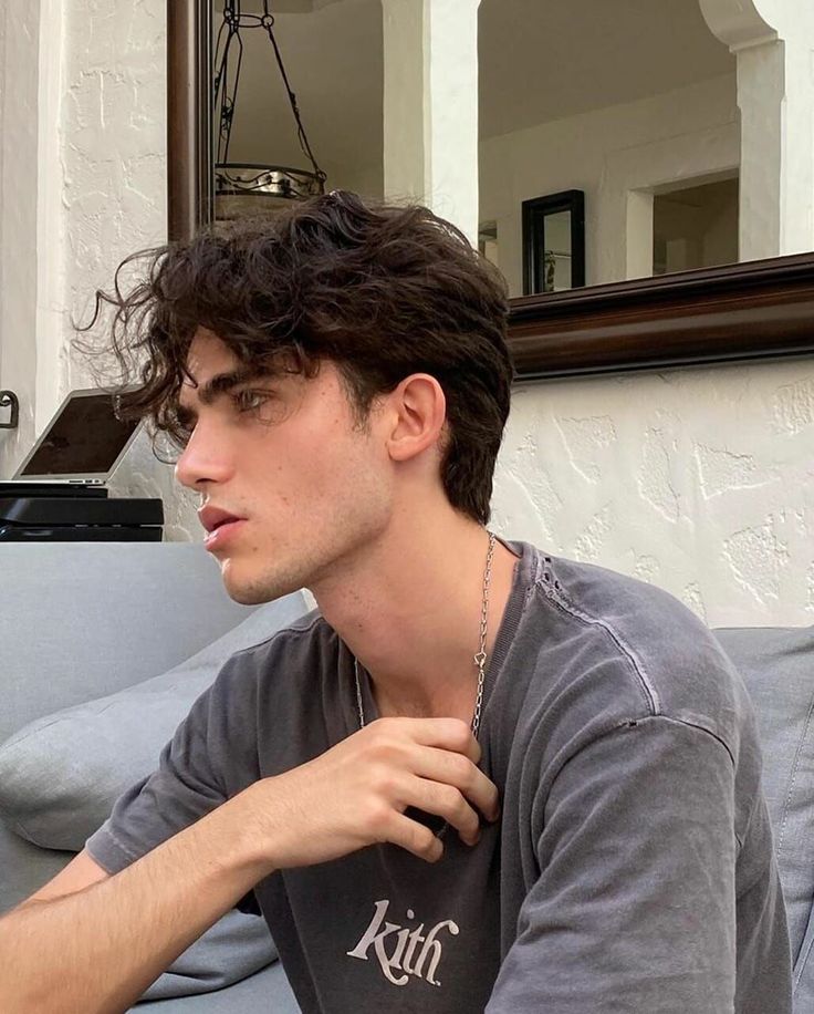 a young man sitting on top of a gray couch next to a window holding a cell phone