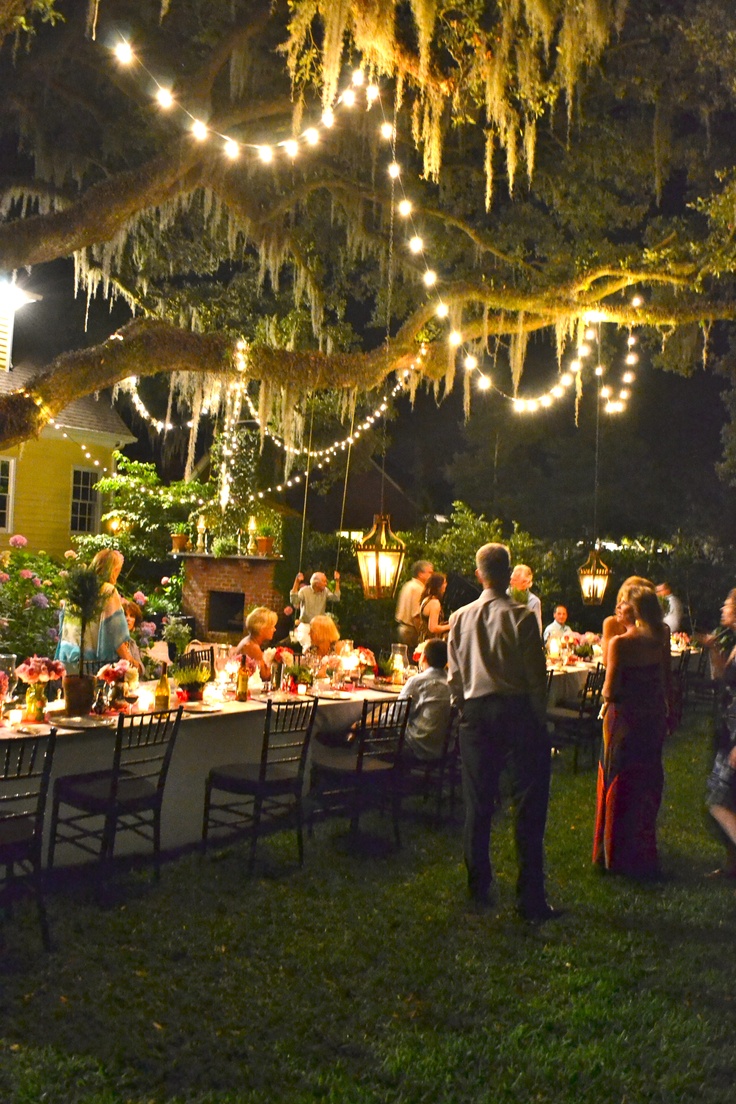 a group of people standing around a dinner table under a tree with lights strung from it