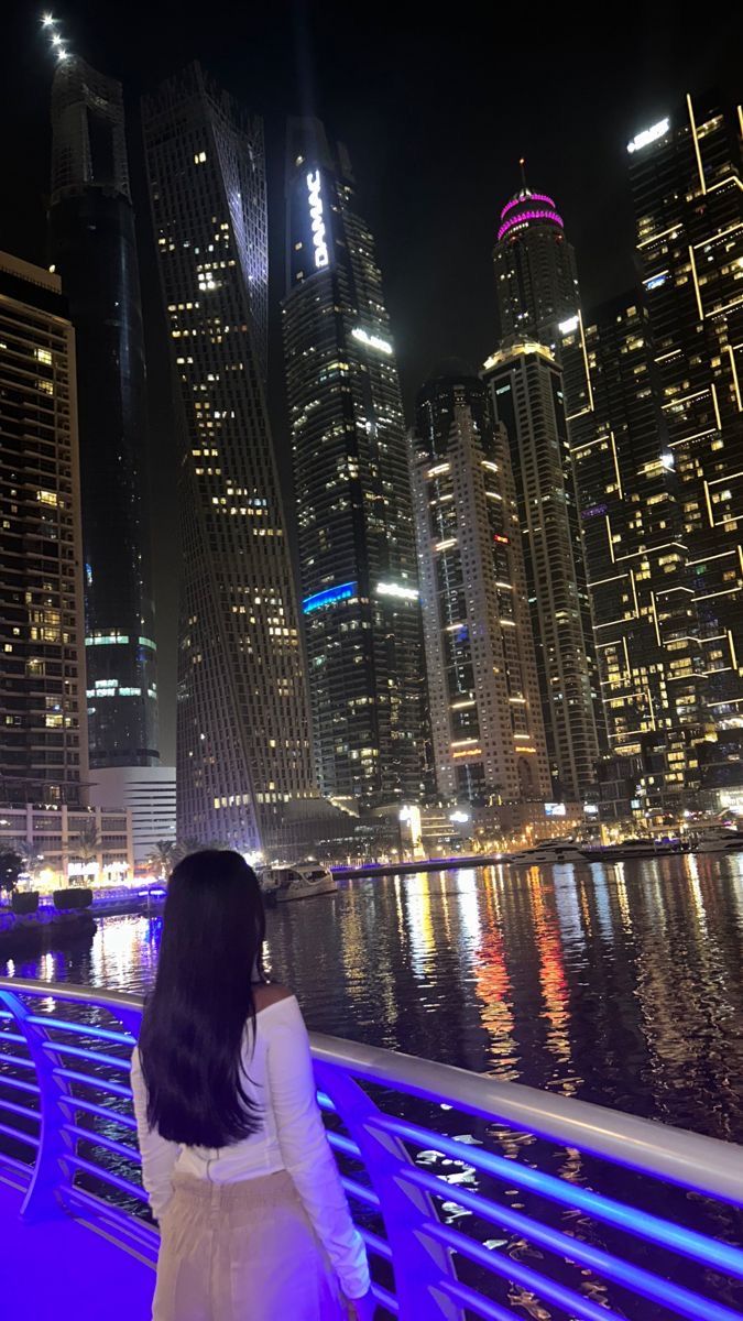 a woman standing on top of a bridge next to the water at night with buildings in the background