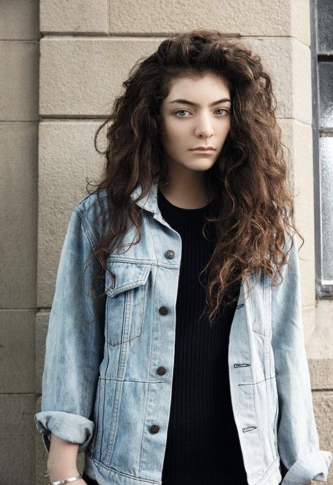 a woman with long hair standing in front of a brick wall wearing a jean jacket