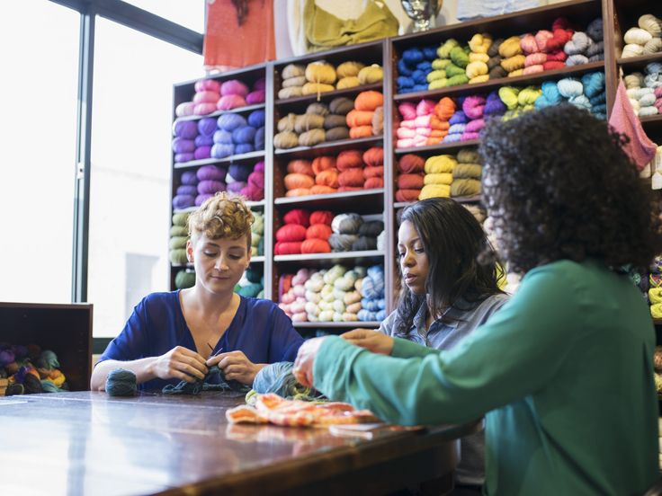 three women sitting at a table in front of a display of crochet yarn