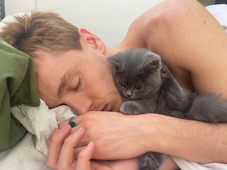 a man laying in bed with a cat sleeping on his chest and holding the kitten