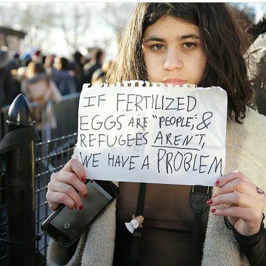 a woman holding up a sign that says if fertiized eggs are people & refugees aren't we have a problem