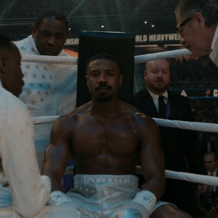 a man sitting in the middle of a boxing ring surrounded by men standing around him