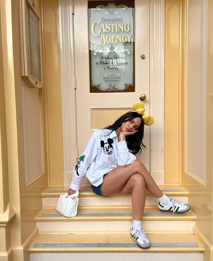 a woman is sitting on the steps in front of a building wearing white sneakers and a mickey mouse shirt