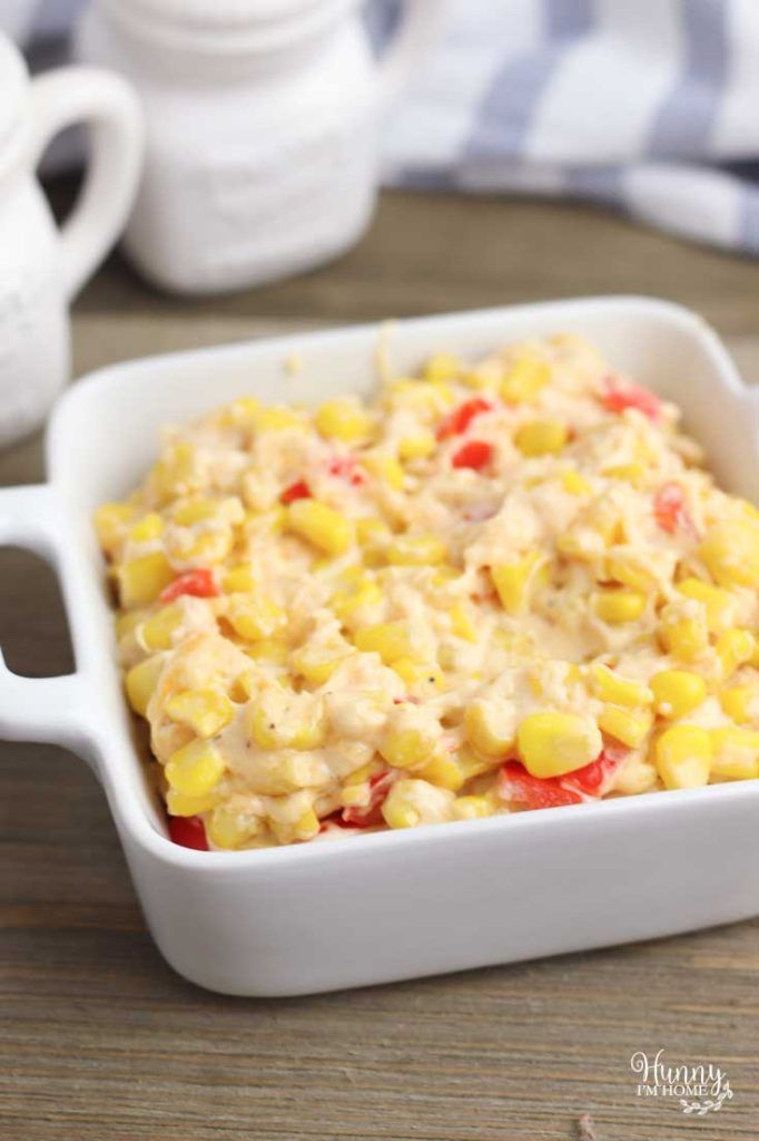 a casserole dish filled with corn on top of a wooden table next to two cups