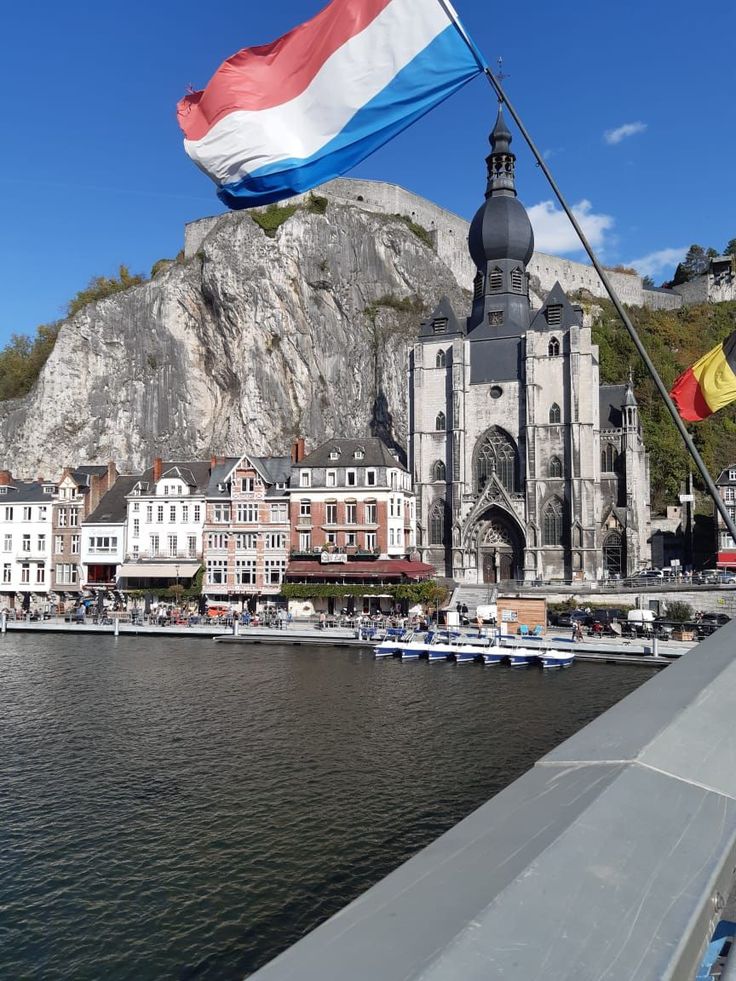 a flag flying in the wind next to a body of water with buildings and mountains in the background