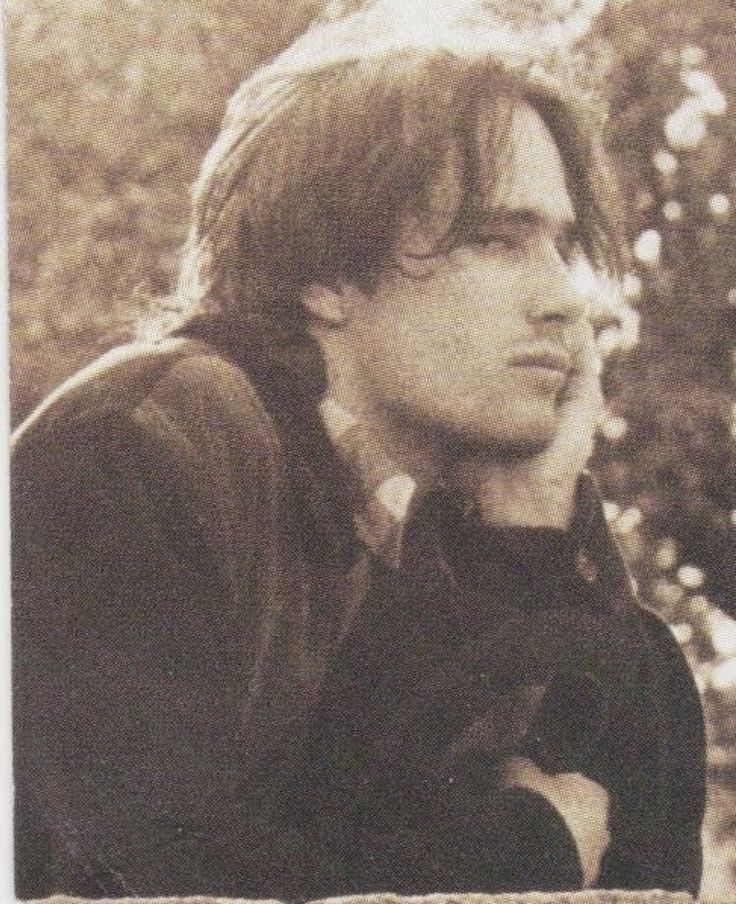 an old photo of a young man with long hair and wearing a black jacket sitting on a bench