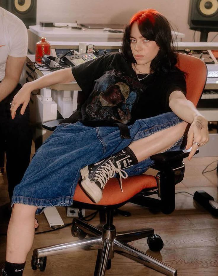 a woman sitting in an office chair with her feet up on the computer desk while another man works behind her