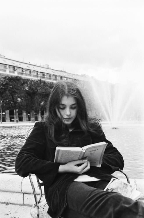 a woman sitting on a bench reading a book in front of a fountain with water spouting behind her