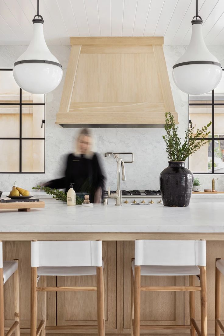 a woman standing in a kitchen next to a counter with chairs and an island top