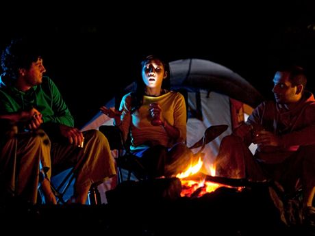 three people sitting around a campfire at night with the lights on and one person looking up