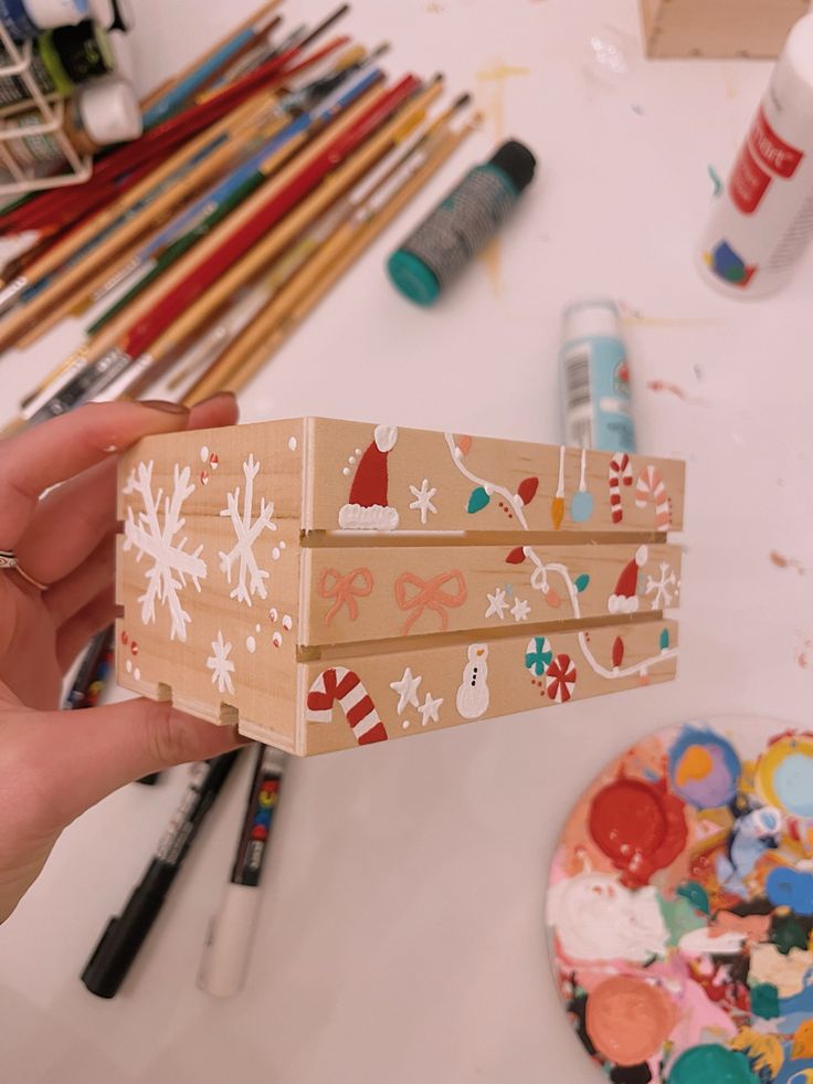 a person holding up a wooden box with christmas decorations on it and paintbrushes nearby