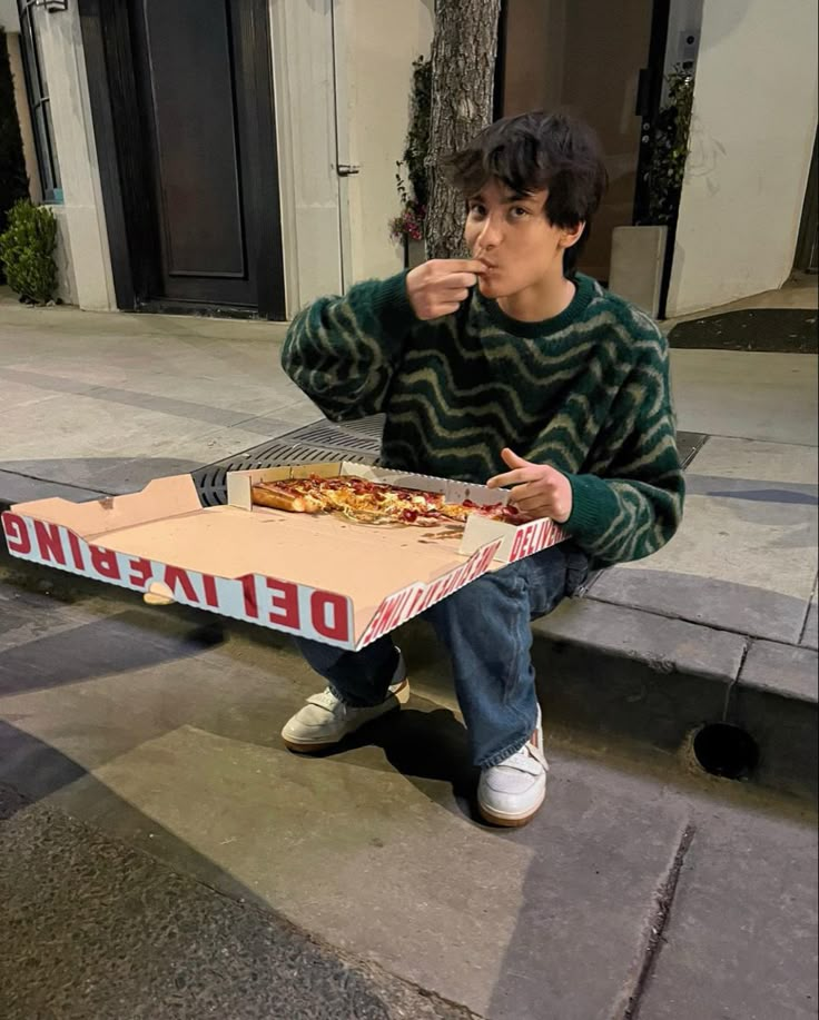 a young man sitting on the ground eating pizza from a box that has been placed in front of him