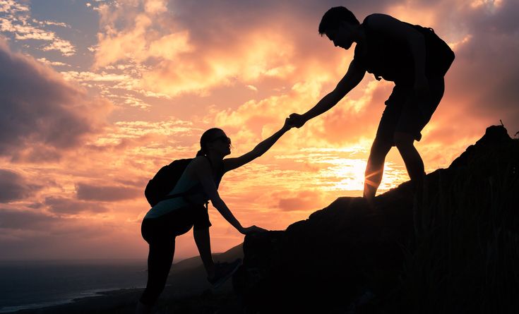 two people are helping each other to climb up a mountain at sunset or sunrise with the sun setting behind them