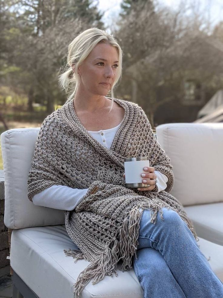 a woman sitting on a white couch holding a coffee cup and wearing a crocheted shawl