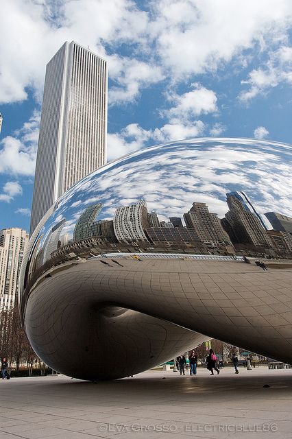 a large metal sculpture in the middle of a city with skyscrapers and clouds behind it